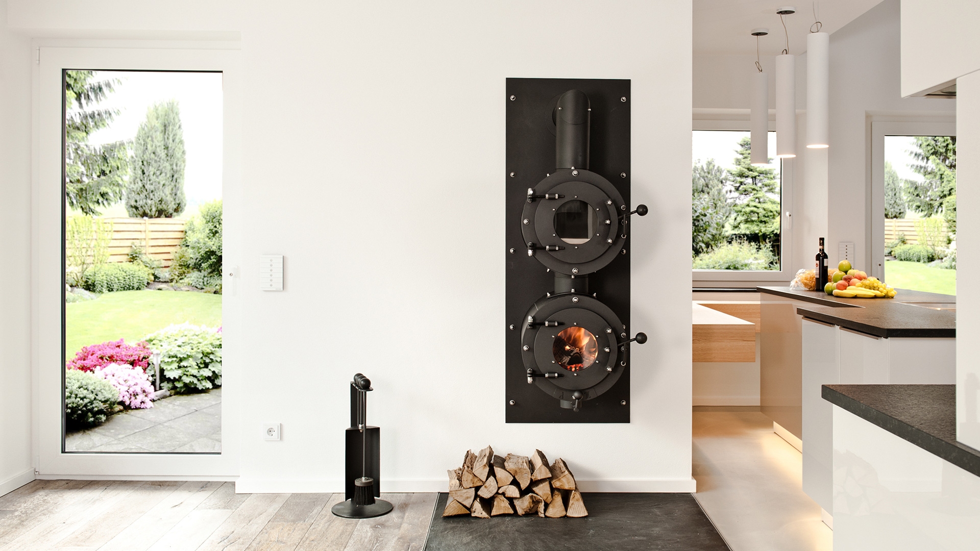 Firetube Ofen mit Garraum darüber, in eine Wohnwand integriert; darunter Holzscheite; rechts Küche, links Terrassentür.