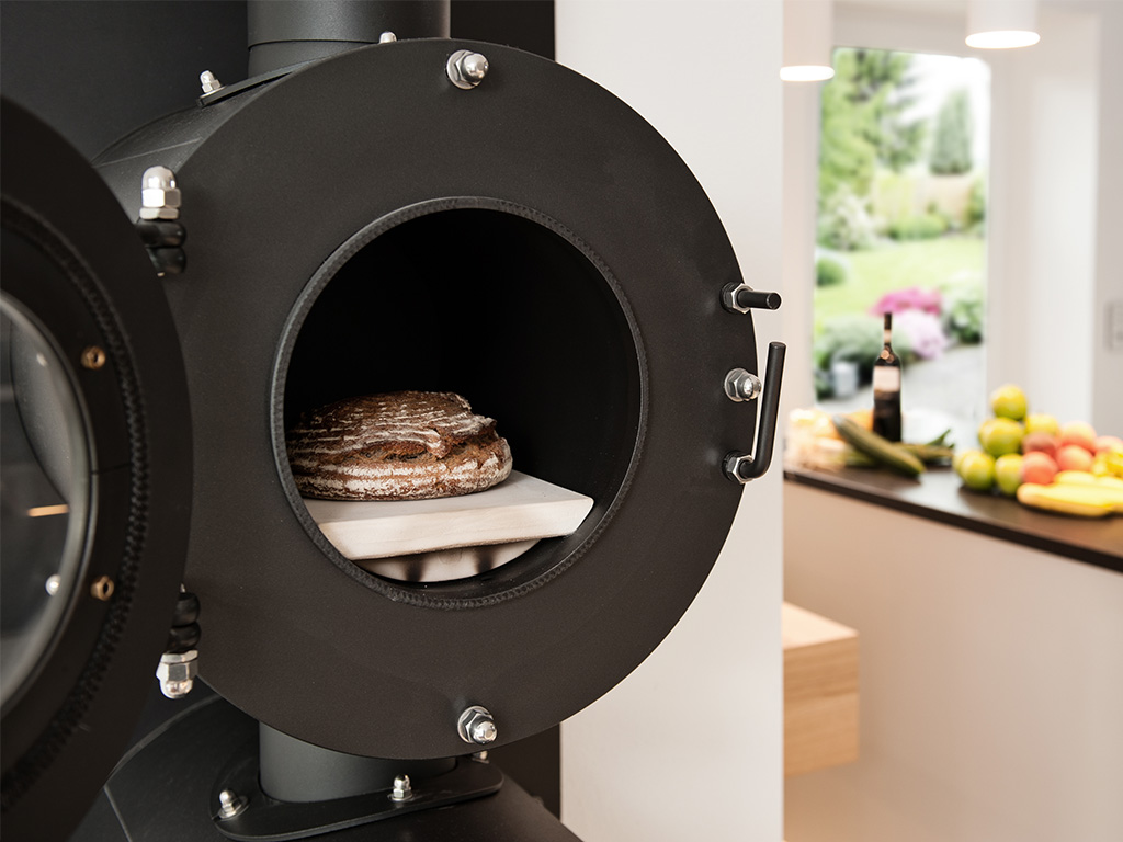 Geöffneter schwarzer firetube Ofen mit Brot im Garraum; im Hintergrund eine Küchenzeile mit Lebensmitteln.