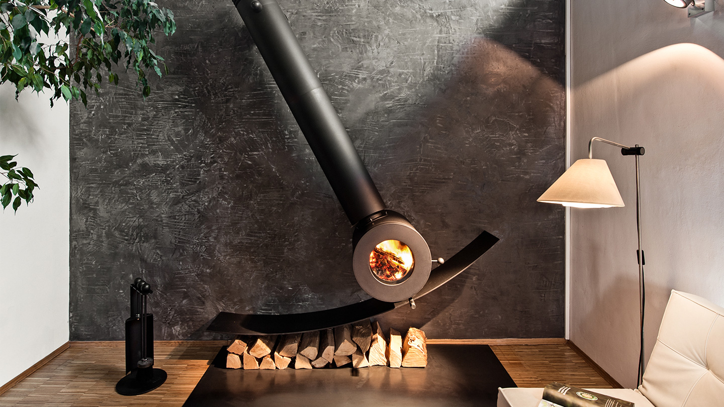 Schräg an der Wand montierter firetube Individualofen mit sichtbarem Feuer; darunter Holzscheite, daneben eine Lampe in wohnlicher Atmosphäre.