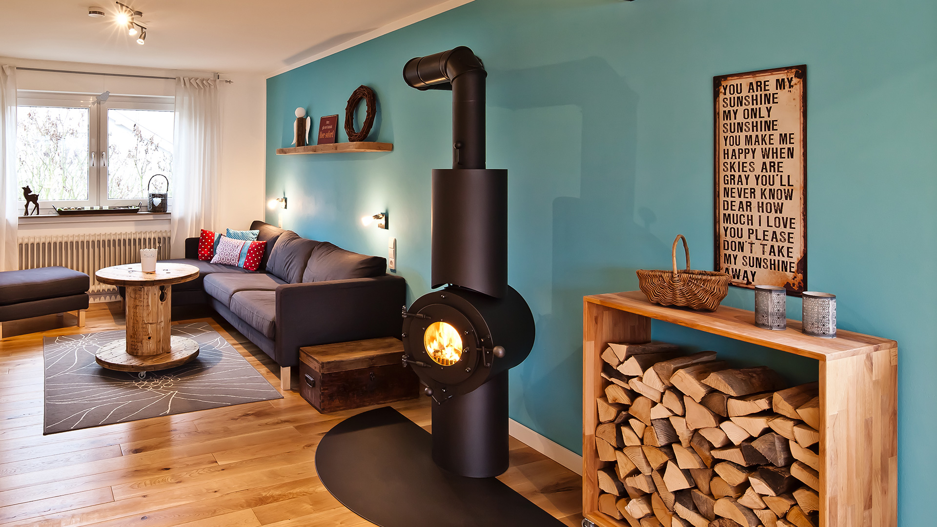 Schwarzer freistehender firetube Ofen mit sichtbarem Feuer in wohnlicher Umgebung; daneben Regal mit gestapelten Holzscheiten, im Hintergrund Sofa und blaue Wand.