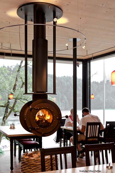 Von der Decke hängender schwarzer firetube Ofen mit sichtbarem Feuer in gastronomischer Umgebung; Gäste und Tische im Hintergrund.