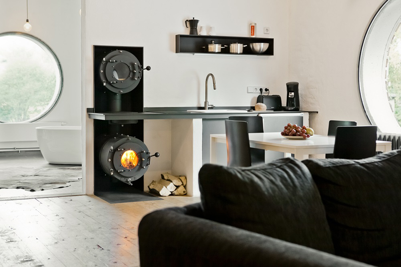 Schwarzer firetube Ofen mit sichtbarem Feuer, in trennende Wand integriert; rechts daneben Küchenzeile, davor Küchentisch und Sofa.