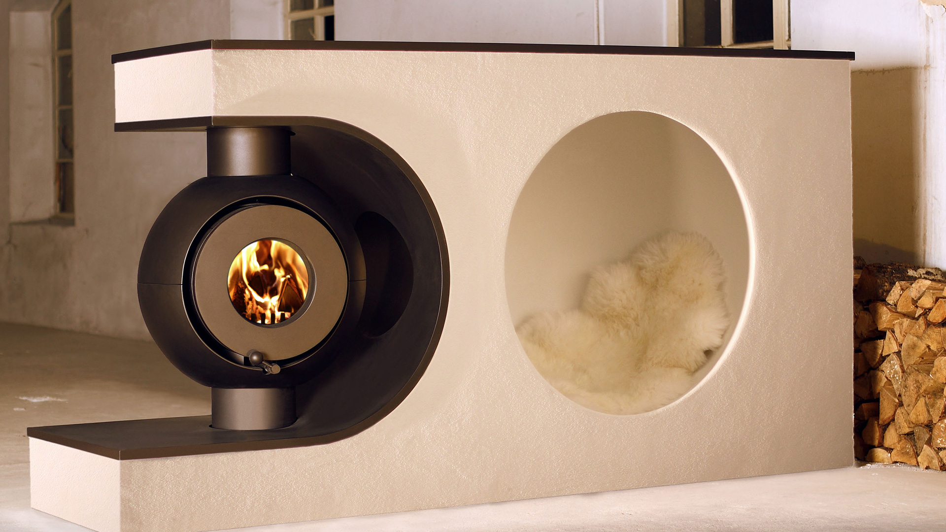 Schwarzer firetube Ofen mit sichtbarem Feuer, wandintegriert neben einer Sitznische mit Fell; Holzscheite im Hintergrund.
