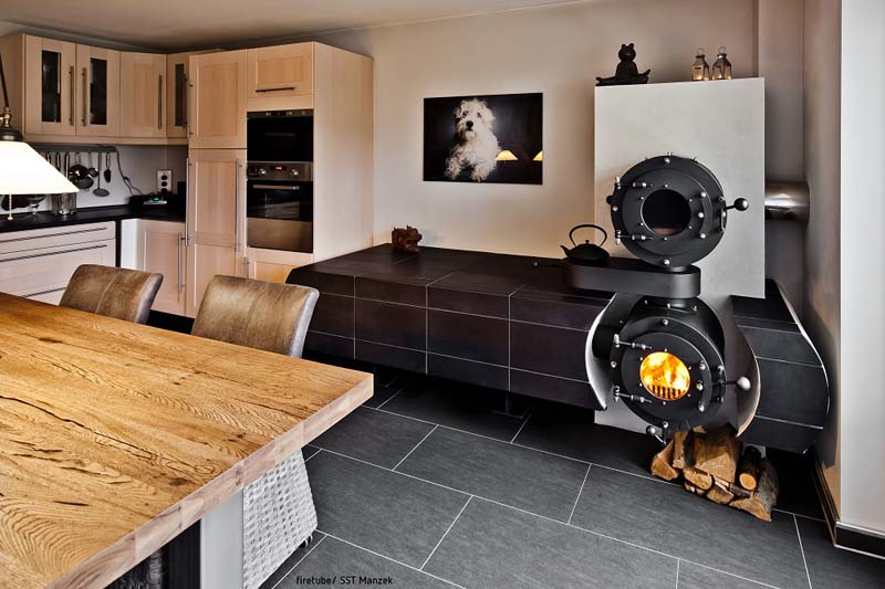 Schwarzer firetube Ofen mit sichtbarem Feuer, Garraum und Hotplate darüber sowie integrierter Sitzfläche in einer Küche; im Hintergrund Küche und Regale.