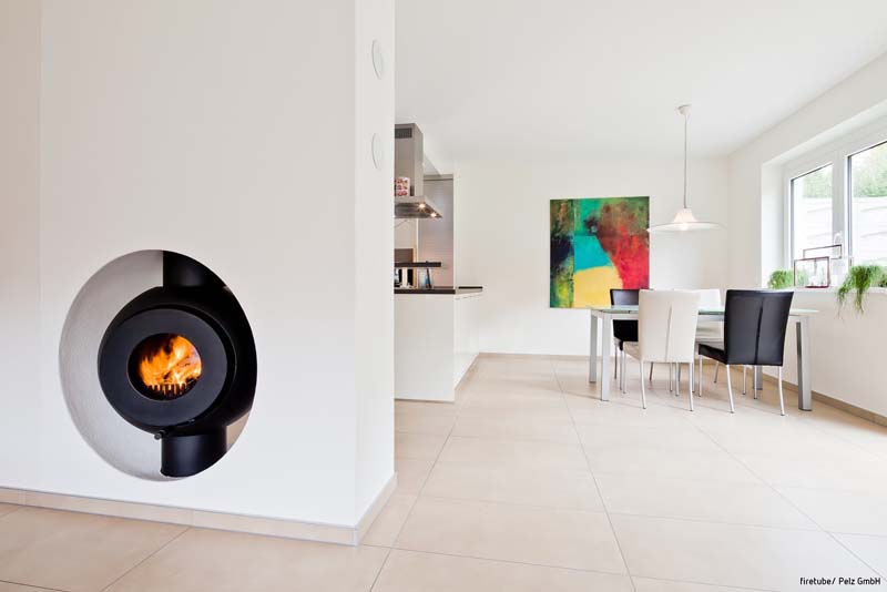 Schwarzer firetube Ofen mit sichtbarem Feuer, wandintegriert; im Hintergrund Wohn- und Essbereich.