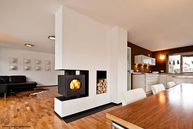 Rechteckiger firetube Ofen mit sichtbarem Feuer, integriert in eine Wohnwand mit Holzfach; Wohn- und Küchenbereich im Umfeld.