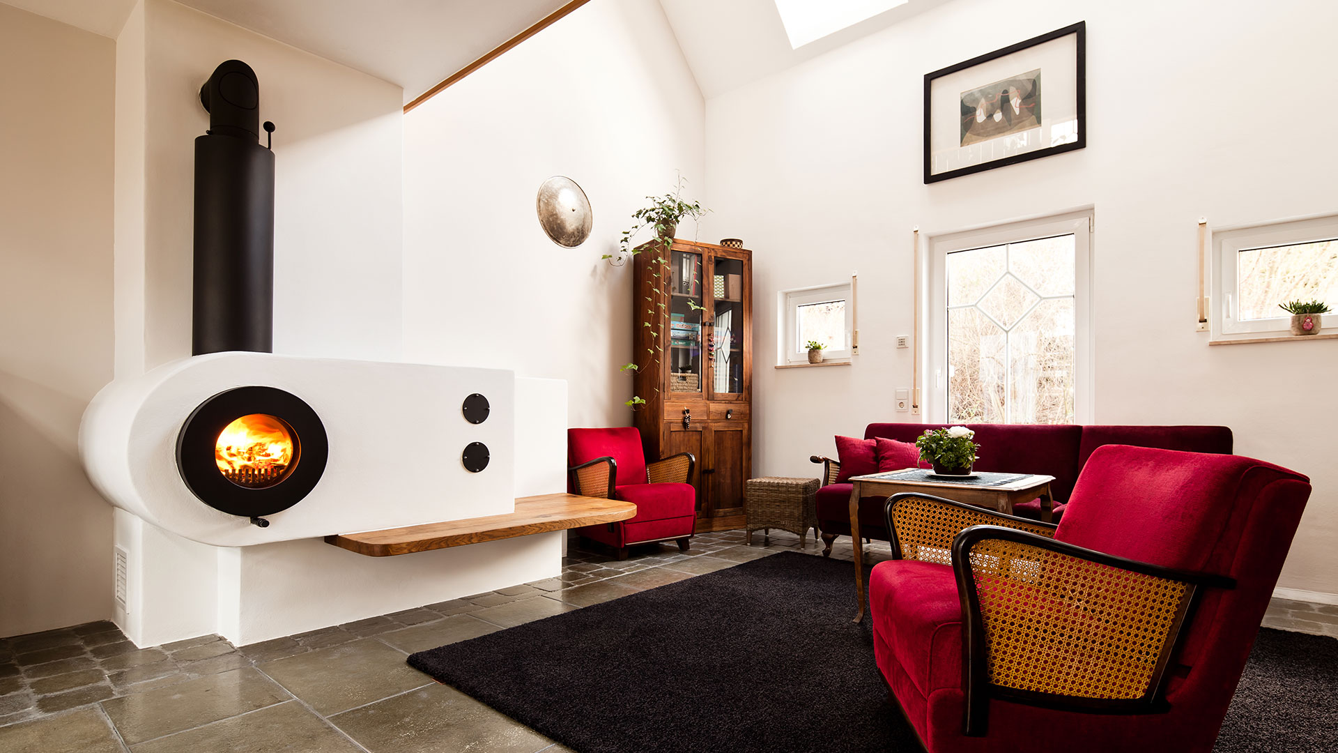 Schwarzer firetube Ofen mit sichtbarem Feuer, in Wohnwand integriert; im Hintergrund Wohnzimmer mit Sofa und Sessel.