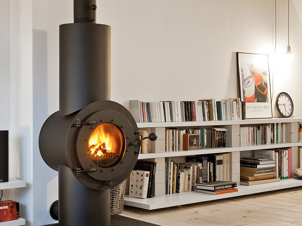 Schwarzer freistehender firetube Ofen mit sichtbarem Feuer in der Brennkammer; im Hintergrund Bücherregal und Lampenlicht.