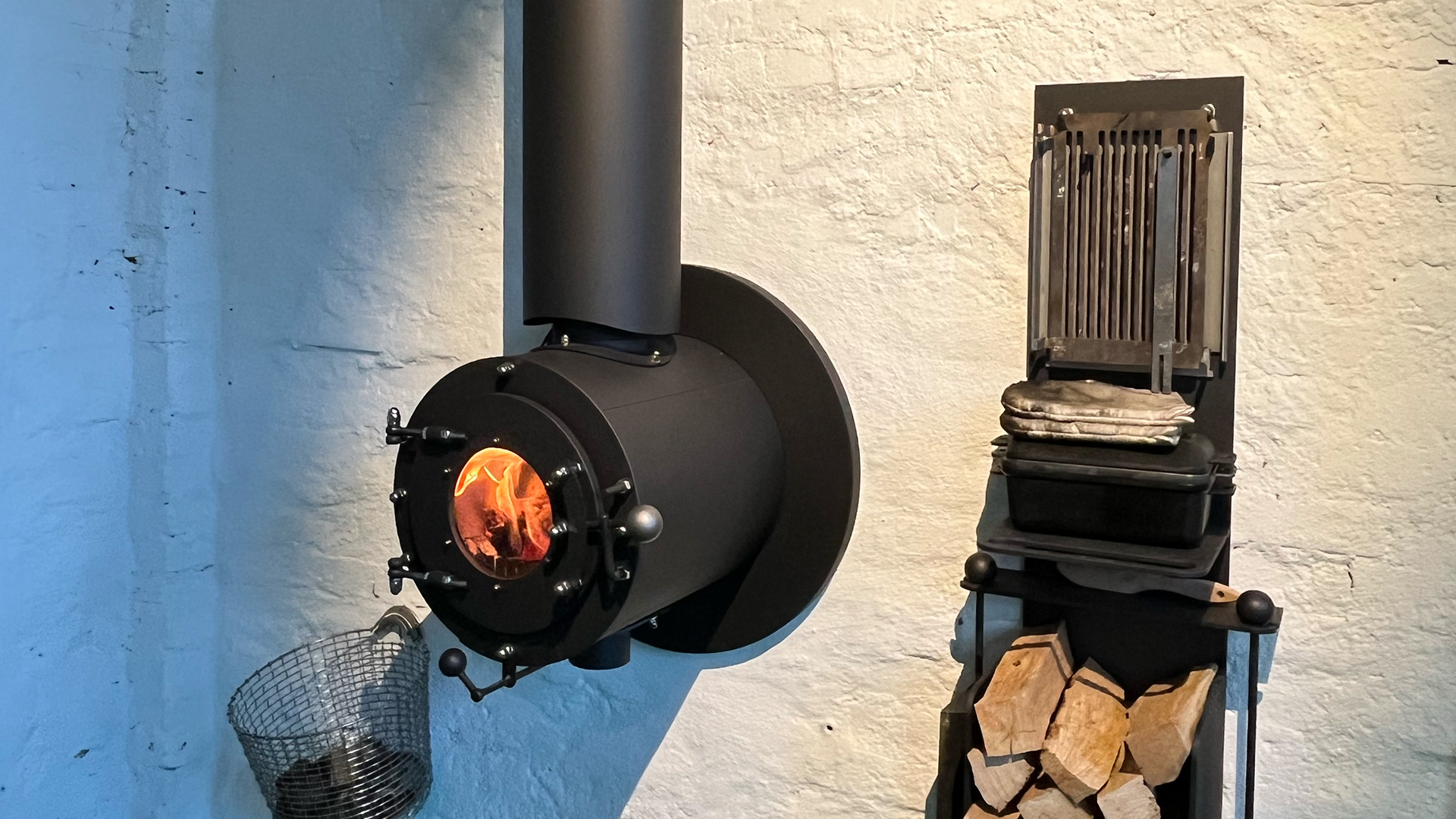 Schwarzer firetube Ofen mit sichtbarem Feuer, in weiße Wand integriert mit herausstehendem Burner; links daneben Holzscheite und rechts ein Behälter mit Kohlen.