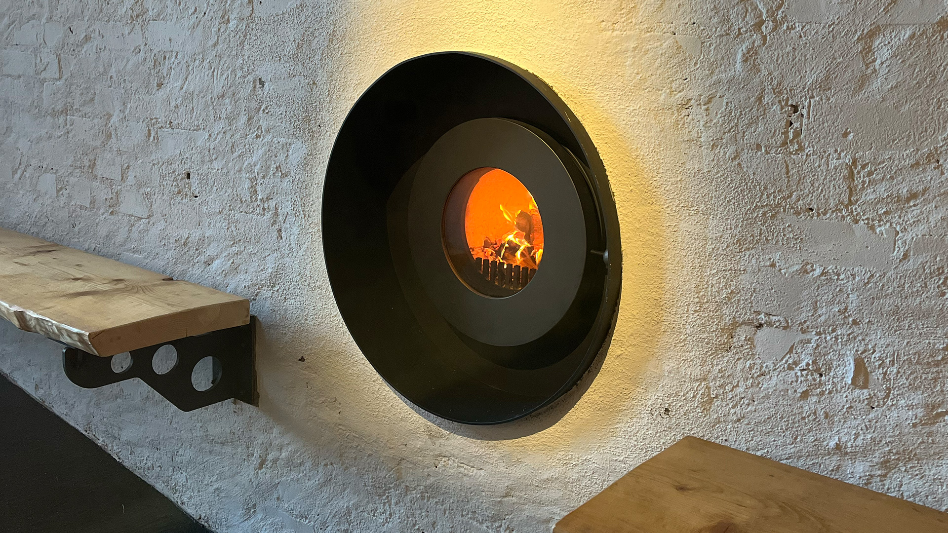 firetube Ofen mit sichtbarem Feuer, vollständig in weiße Wand integriert; rechts und links Holzregale befestigt.