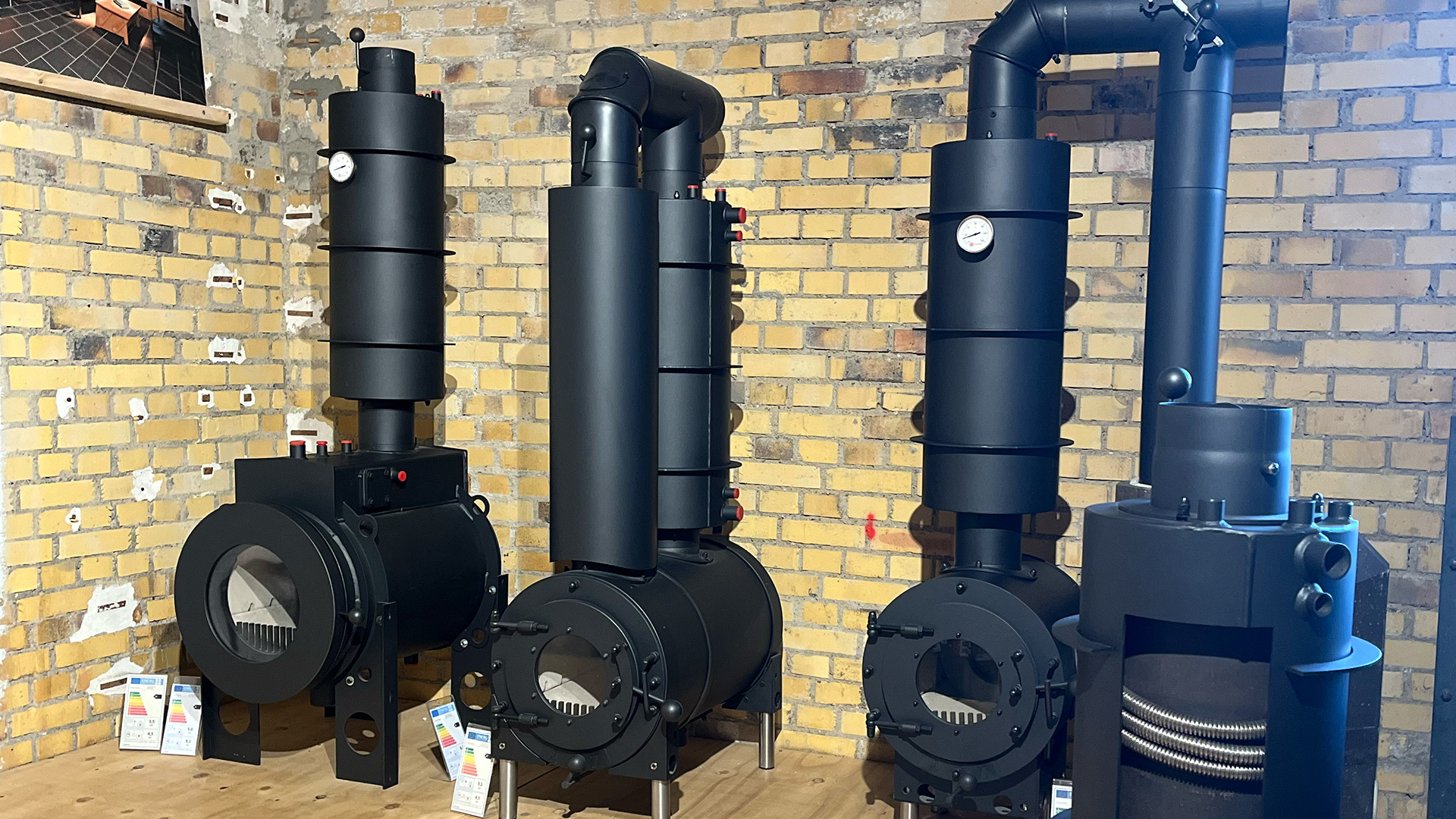 Drei schwarze firetube Öfen nebeneinander in unterschiedlichen Ausführungen, vor einer Steinwand im Hintergrund.