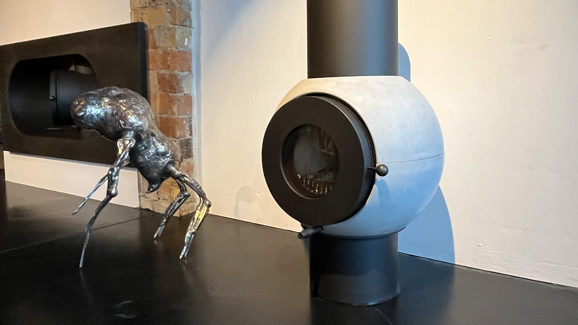Grau-schwarzer firetube Ofen freistehend im Raum, daneben eine Skulptur; im Hintergrund in Wand integrierter weiterer firetube Ofen.