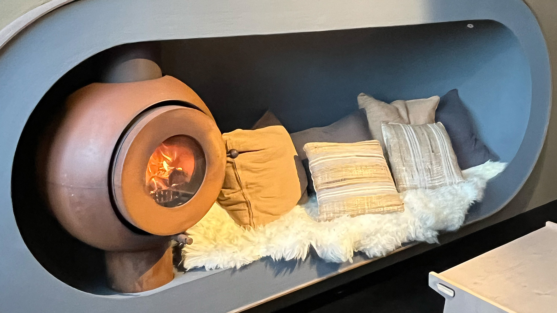 Firetube Ofen mit sichtbarem Feuer im rustikalen Look, in einer Sitzkuhle mit Kissen und Felldecke.