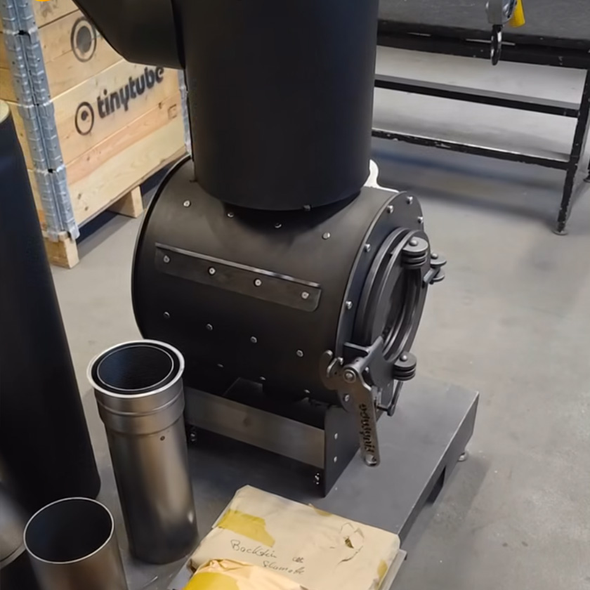 Schwarzer tinytube Ofen in Seitenansicht; daneben zwei Rohre, im Hintergrund Holzkisten mit Aufschrift „tinytube“.