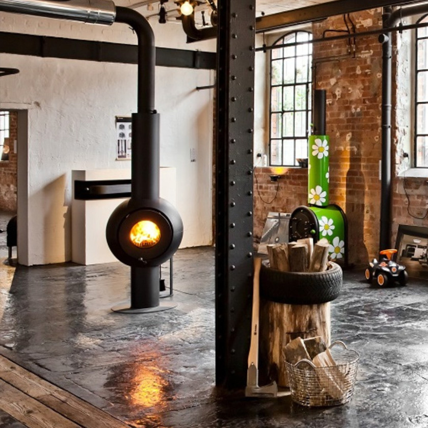 Schwarzer freistehender firetube Ofen mit sichtbarem Feuer in rustikaler Umgebung; davor Holzbehälter mit Holzscheiten und Axt, dahinter ein grün lackierter firetube Ofen mit Blumenmotiv.