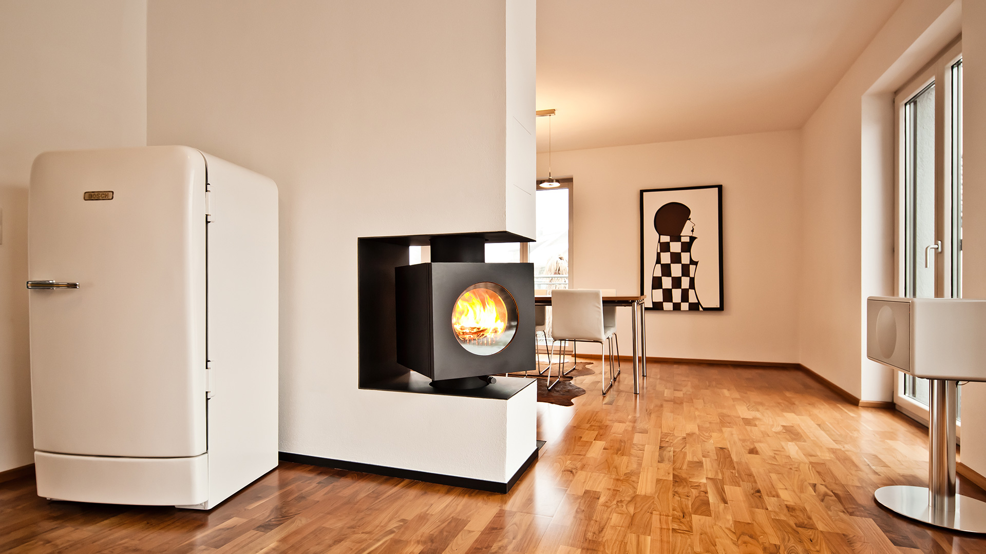 Schwarzer rechteckiger firetube Ofen mit sichtbarem Feuer, integriert in eine trennende Wohnwand; im Hintergrund Wohnbereich.