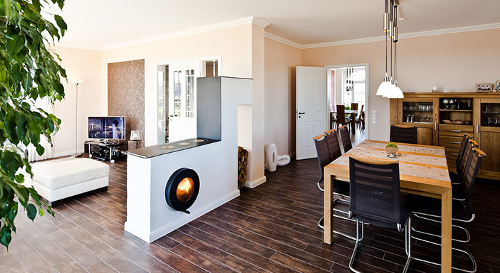 Firetube Ofen mit sichtbarem Feuer, in Küchenzeile integriert; darauf Kochplatten, dahinter gestapelte Holzscheite, drumherum Küchentisch und Wohnbereich.