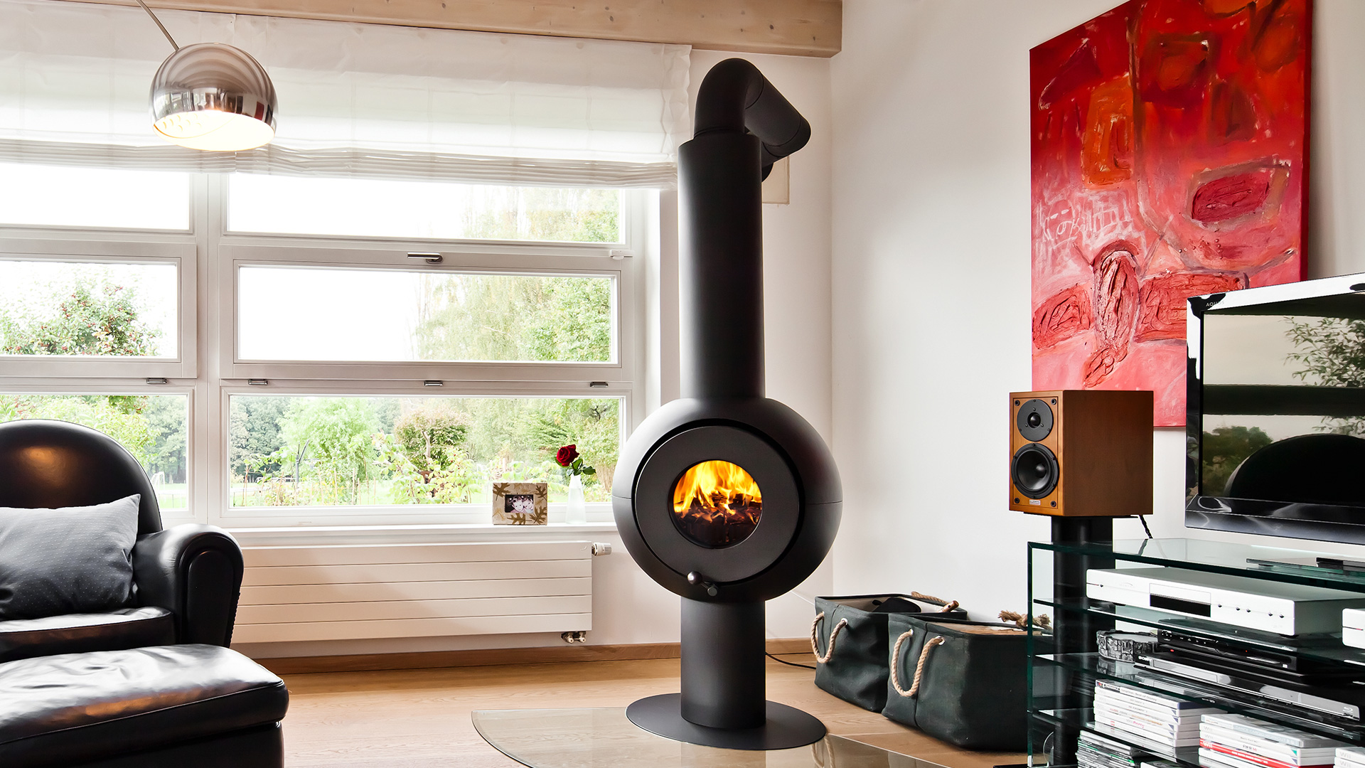 Schwarzer freistehender firetube Ofen mit sichtbarem Feuer in wohnlicher Umgebung; im Hintergrund Fenster mit Blick auf Wald.
