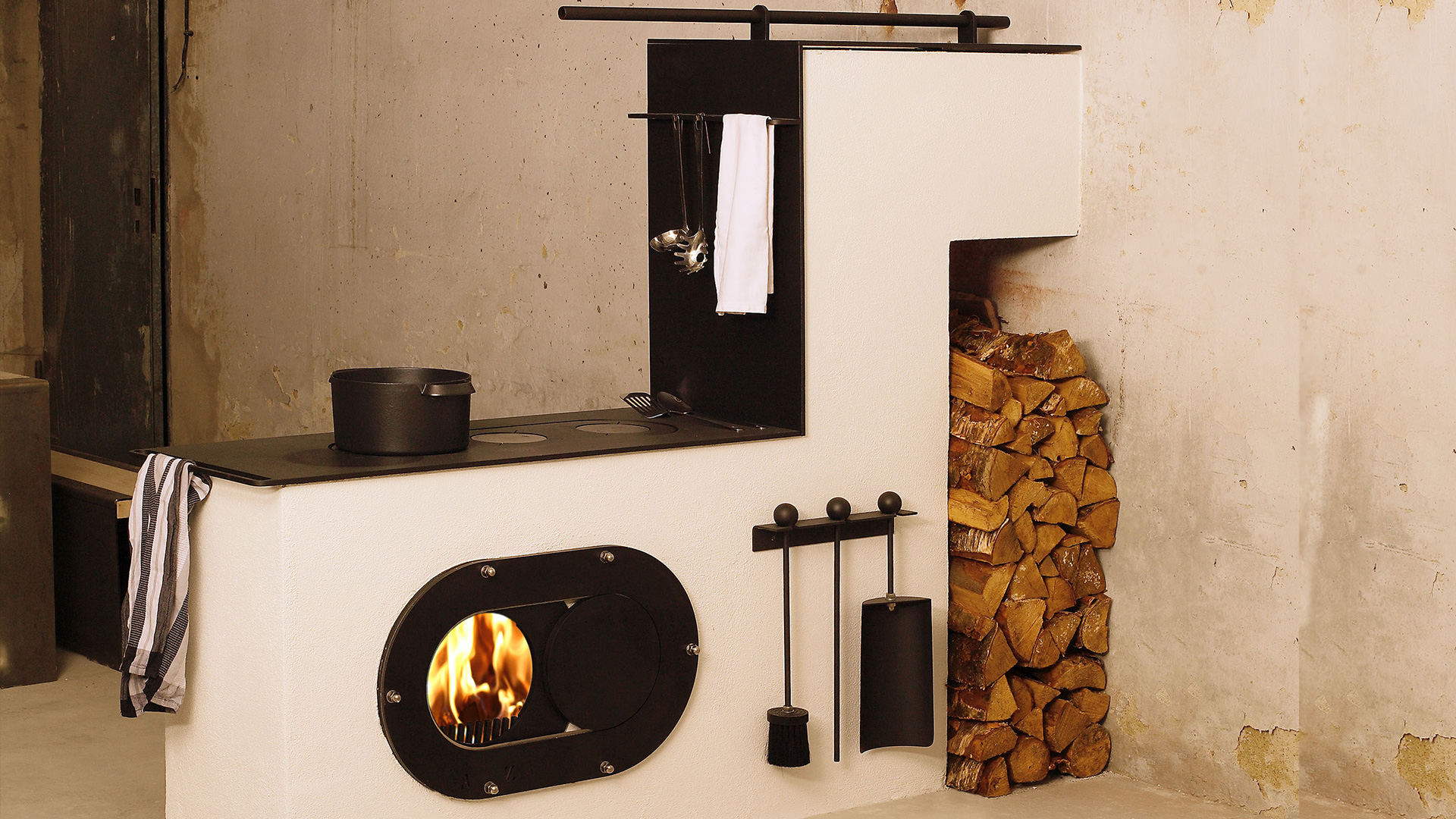 Schwarzer firetube Ofen mit sichtbarem Feuer, eingebaut in eine trennende flache Wand mit Kochplatten darauf; dahinter Holzscheite.