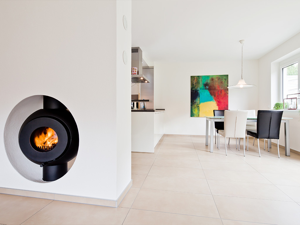 Schwarzer firetube Ofen mit sichtbarem Feuer, wandintegriert; im Hintergrund Wohn- und Essbereich.