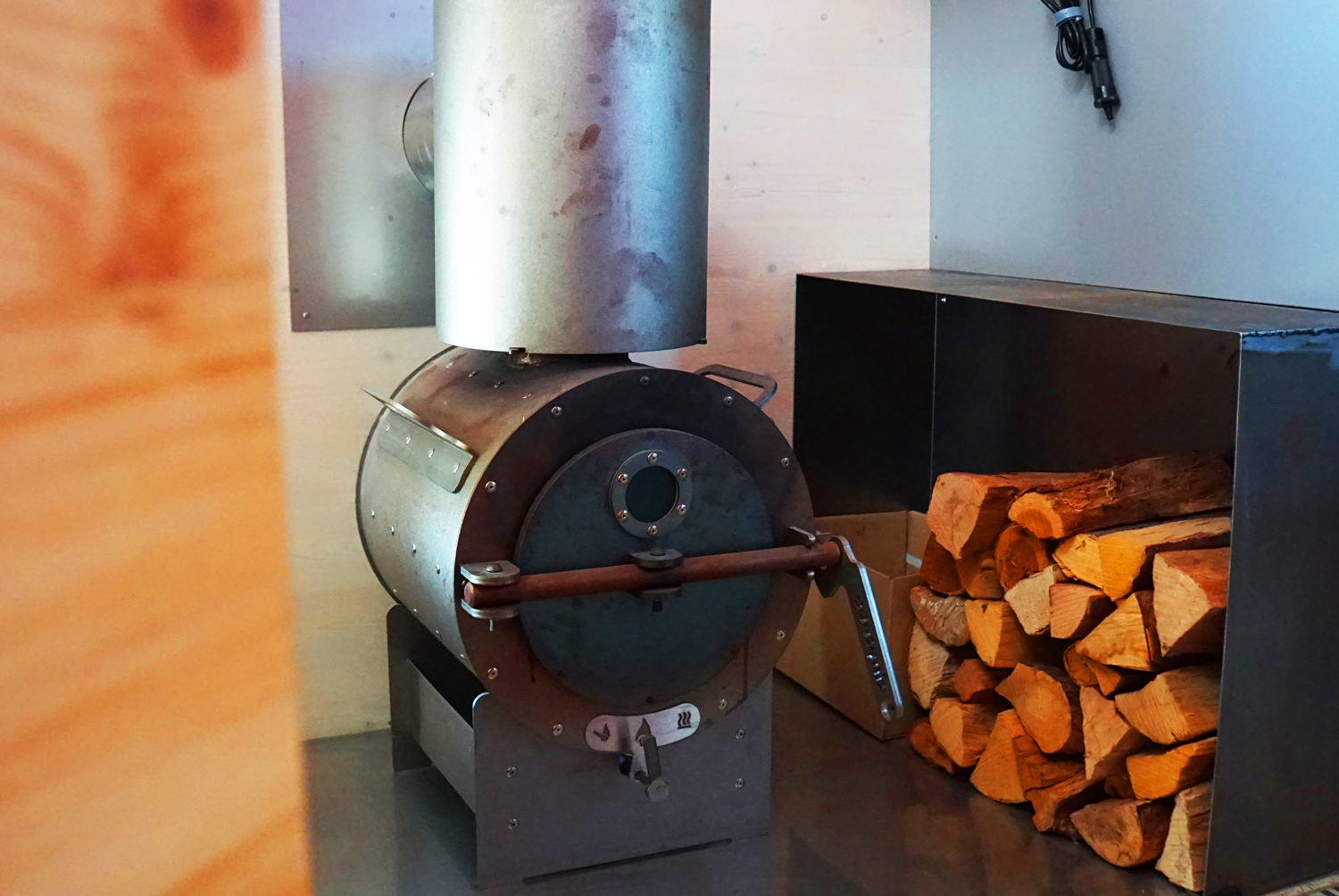tinytube Ofen freistehend auf dem Boden, daneben gestapelte Holzscheite in einer Halterung; Abzug und Wandflächen im Hintergrund.