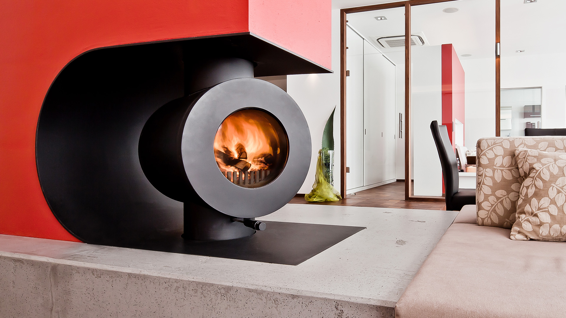 In eine Wohnlandschaft integrierter schwarzer firetube Speicherofen mit sichtbarem Feuer in rotem Wandausschnitt mit Sitzfläche; im Hintergrund ein Raum mit Glaswänden.